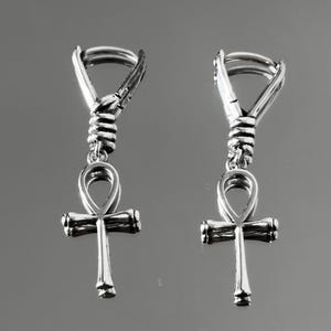 May include: A pair of silver-toned metal earrings featuring an ankh symbol pendant suspended from a coiled wire design. Each earring includes a hinged hoop closure at the top, with the ankh cross dangling below the textured, wrapped metal detail.