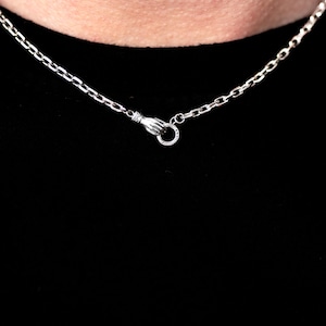 May include: A silver-toned chain necklace featuring a clasp designed as a hand holding a small circular ring. The metal links are uniform and connected to the hand-shaped fastener, which secures the chain around the neck area.