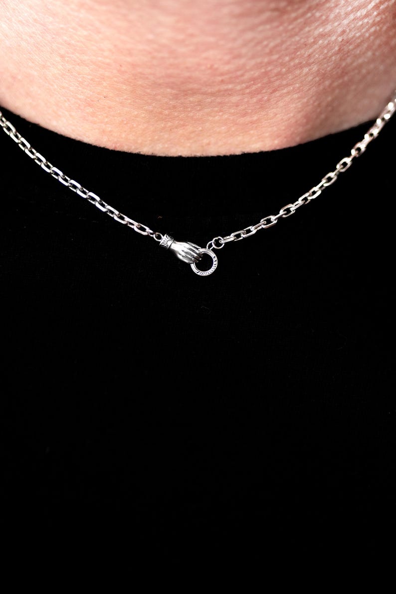 May include: A silver-toned chain necklace featuring a clasp designed as a hand holding a small circular ring. The metal links are uniform and connected to the hand-shaped fastener, which secures the chain around the neck area.