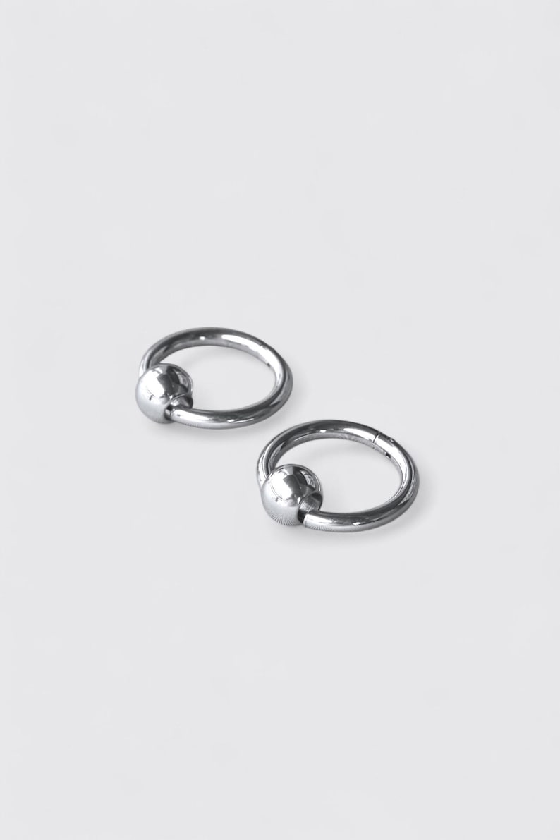 May include: Two silver-toned circular rings, each featuring a smooth, polished spherical bead attached to the hoop. These metallic jewelry pieces have a reflective, high-shine finish and a simple, continuous circular design with a secure closure.