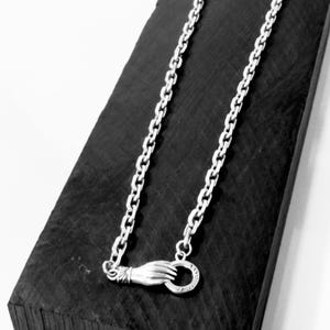 May include: A silver-toned metal chain necklace featuring a clasp designed as a hand holding a circular ring. The chain consists of uniform oval links, and the hand-shaped clasp is positioned at the bottom of the draped necklace.