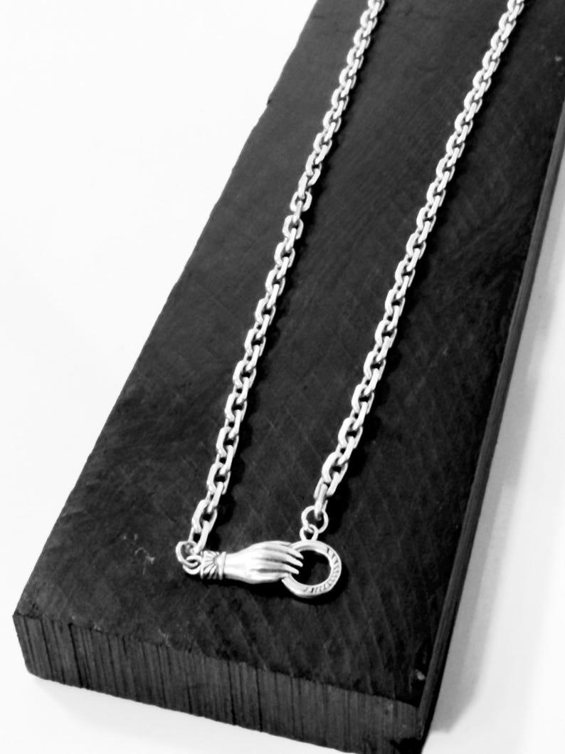 May include: A silver-toned metal chain necklace featuring a clasp designed as a hand holding a circular ring. The chain consists of uniform oval links, and the hand-shaped clasp is positioned at the bottom of the draped necklace.