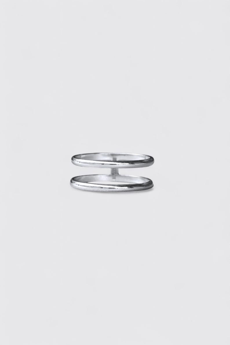 K&ouml;nnte beinhalten: Ein silberfarbener Ring mit zwei parallelen B&auml;ndern, die durch einen vertikalen Steg in der Mitte verbunden sind. Das Metall hat eine glatte, polierte Oberfl&auml;che mit einem reflektierenden Glanz, was eine schlichte Struktur f&uuml;r diesen Ring ergibt.