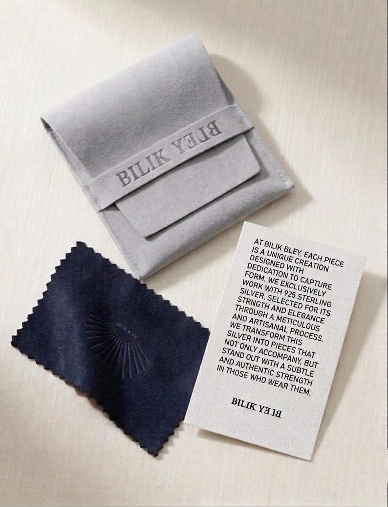 May include: A light gray velvet pouch with the text BILIK YELIB, a dark navy blue polishing cloth with a sunburst emblem, and a white rectangular card featuring printed text about 925 sterling silver and the brand name BILIK YELIB.