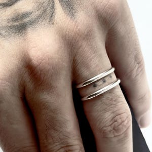 K&ouml;nnte beinhalten: Ein Doppelring bestehend aus zwei glatten, polierten silberfarbenen Metallringen, die durch einen kleinen vertikalen Steg verbunden sind und einen offenen Raum zwischen den B&auml;ndern bilden. Das Design hat eine klare, metallische Oberfl&auml;che.