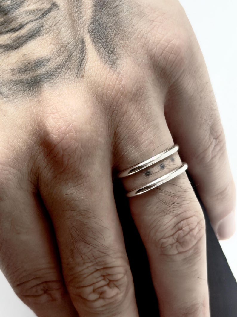 K&ouml;nnte beinhalten: Ein Doppelring bestehend aus zwei glatten, polierten silberfarbenen Metallringen, die durch einen kleinen vertikalen Steg verbunden sind und einen offenen Raum zwischen den B&auml;ndern bilden. Das Design hat eine klare, metallische Oberfl&auml;che.