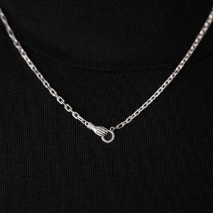 May include: A silver-toned metal chain necklace featuring a clasp designed as a small hand holding a circular ring. The chain links are uniform and oval-shaped, resting against a solid black fabric background to highlight the metallic finish.