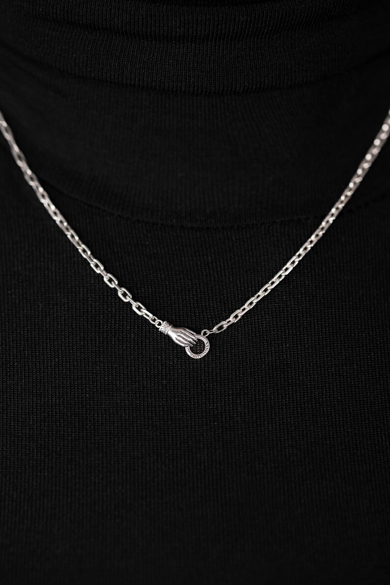 May include: A silver-toned metal chain necklace featuring a clasp designed as a small hand holding a circular ring. The chain links are uniform and oval-shaped, resting against a solid black fabric background to highlight the metallic finish.
