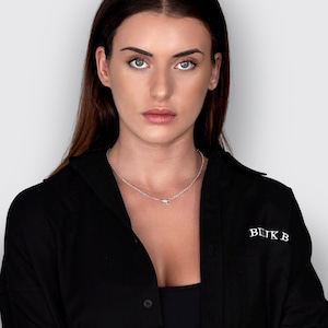 May include: A black button-down shirt featuring the white text BLIK B embroidered on the chest. The shirt is worn with a simple silver-toned chain necklace that rests against the collarbone, completing the look with a clean, dark aesthetic.