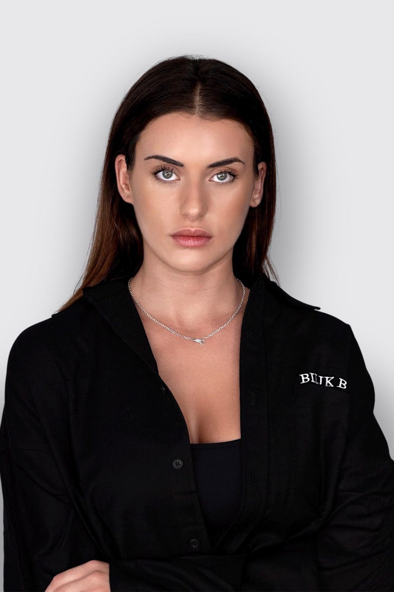 May include: A black button-down shirt featuring the white text BLIK B embroidered on the chest. The shirt is worn with a simple silver-toned chain necklace that rests against the collarbone, completing the look with a clean, dark aesthetic.