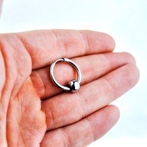 May include: A silver-toned circular hoop earring featuring a single polished spherical bead attached to the ring. The hoop has a hinged closure mechanism visible at the top, and the entire piece is constructed from a smooth, reflective metallic material.