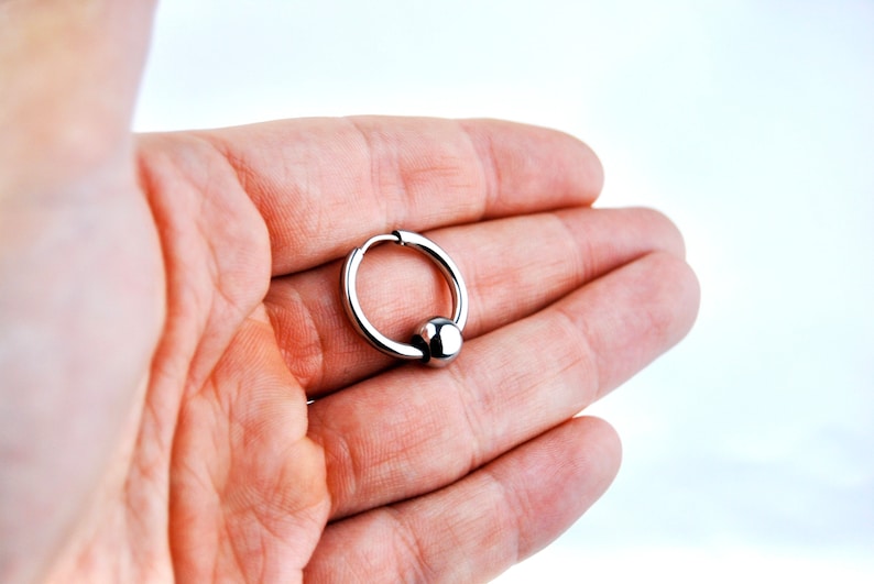 May include: A silver-toned circular hoop earring featuring a single polished spherical bead attached to the ring. The hoop has a hinged closure mechanism visible at the top, and the entire piece is constructed from a smooth, reflective metallic material.