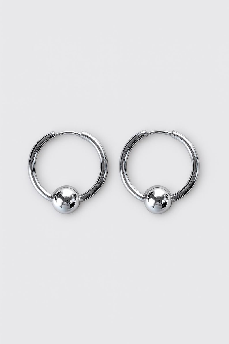 May include: A pair of silver-toned hoop earrings, each featuring a smooth, polished spherical bead centered on the bottom of the circular wire hoop. The earrings have a reflective metallic finish and a simple, clean design with a hinged closure mechanism.