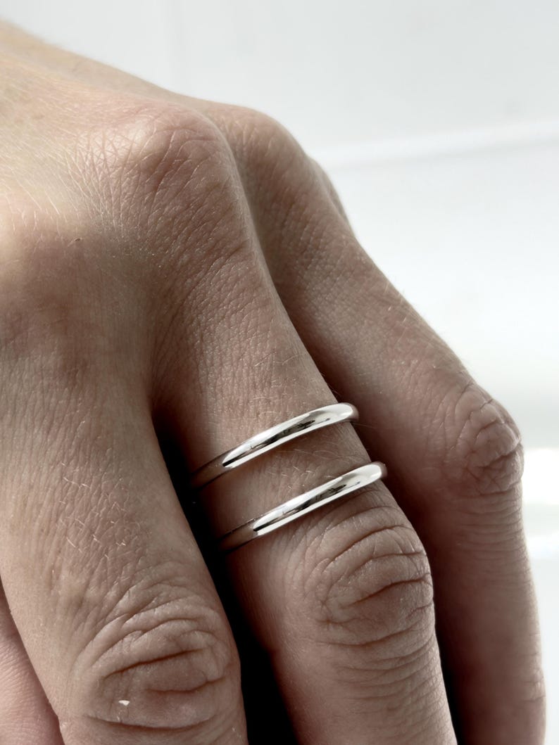 K&ouml;nnte beinhalten: Zwei polierte Silberringe an einem Finger. Jeder Ring hat ein glattes, abgerundetes Profil mit einer hochgl&auml;nzenden metallischen Oberfl&auml;che. Die Ringe sind schlicht, einfach und von gleichm&auml;&szlig;iger Dicke, was ein klares und reflektierendes Erscheinungsbild erzeugt.