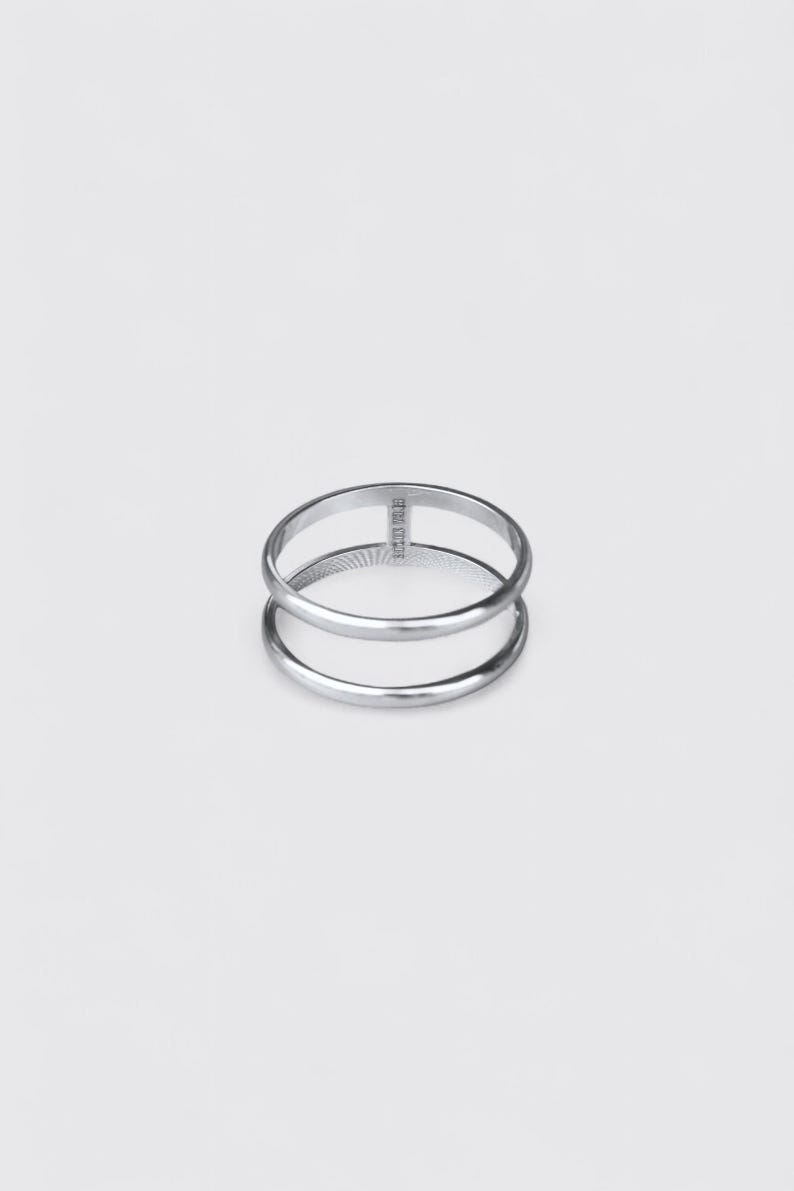 K&ouml;nnte beinhalten: Ein silberfarbener Metallring mit einem Doppelband-Design. Die beiden B&auml;nder sind an einer Seite durch einen kleinen vertikalen Steg miteinander verbunden. Die Oberfl&auml;che des Rings ist glatt und poliert, und das Metall wirkt im Licht reflektierend.