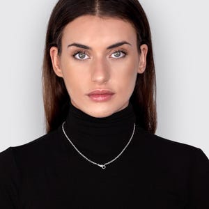 May include: A simple silver chain necklace featuring small, uniform oval links and a secure lobster claw clasp, worn around the neck against a black turtleneck shirt. The metallic finish of the silver chain provides a bright contrast.