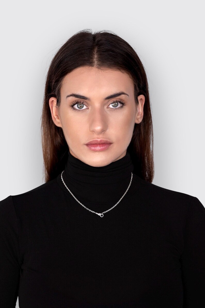 May include: A simple silver chain necklace featuring small, uniform oval links and a secure lobster claw clasp, worn around the neck against a black turtleneck shirt. The metallic finish of the silver chain provides a bright contrast.