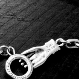 May include: A silver-toned metal necklace clasp shaped like a hand holding a circular ring. The hand features the engraved text S925 and ER-1862. The clasp is attached to a chain link, showcasing a detailed metallic finish and design.