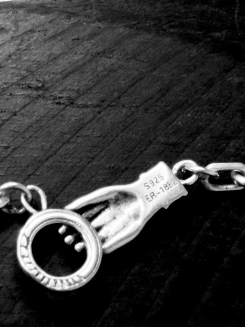 May include: A silver-toned metal necklace clasp shaped like a hand holding a circular ring. The hand features the engraved text S925 and ER-1862. The clasp is attached to a chain link, showcasing a detailed metallic finish and design.