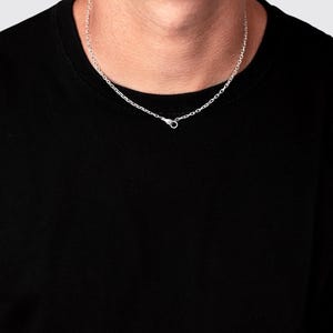 May include: A silver chain necklace featuring a small, detailed hand-shaped clasp at the front. The necklace is worn around the neck against a plain black shirt, showcasing the metallic links and the unique design of the closure mechanism.