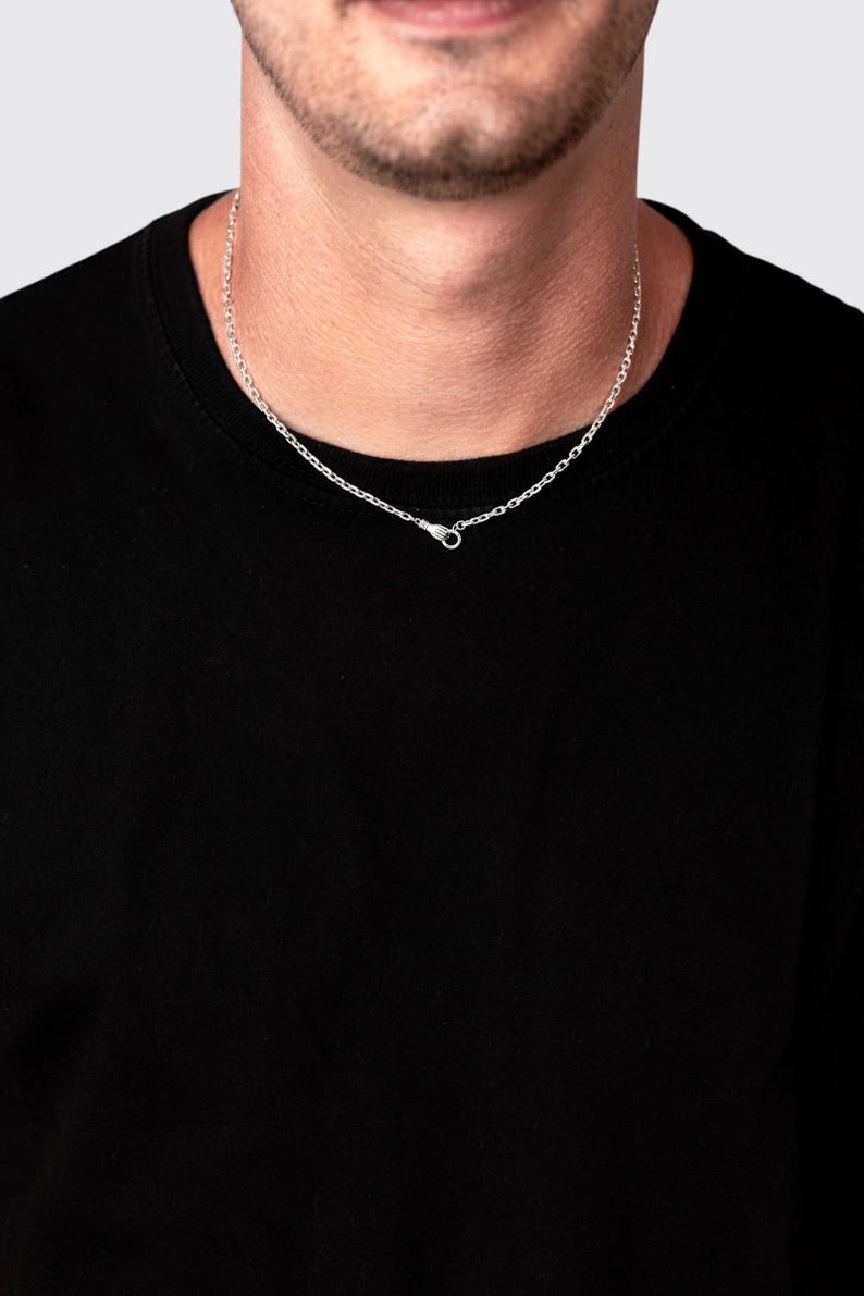 May include: A silver chain necklace featuring a small, detailed hand-shaped clasp at the front. The necklace is worn around the neck against a plain black shirt, showcasing the metallic links and the unique design of the closure mechanism.