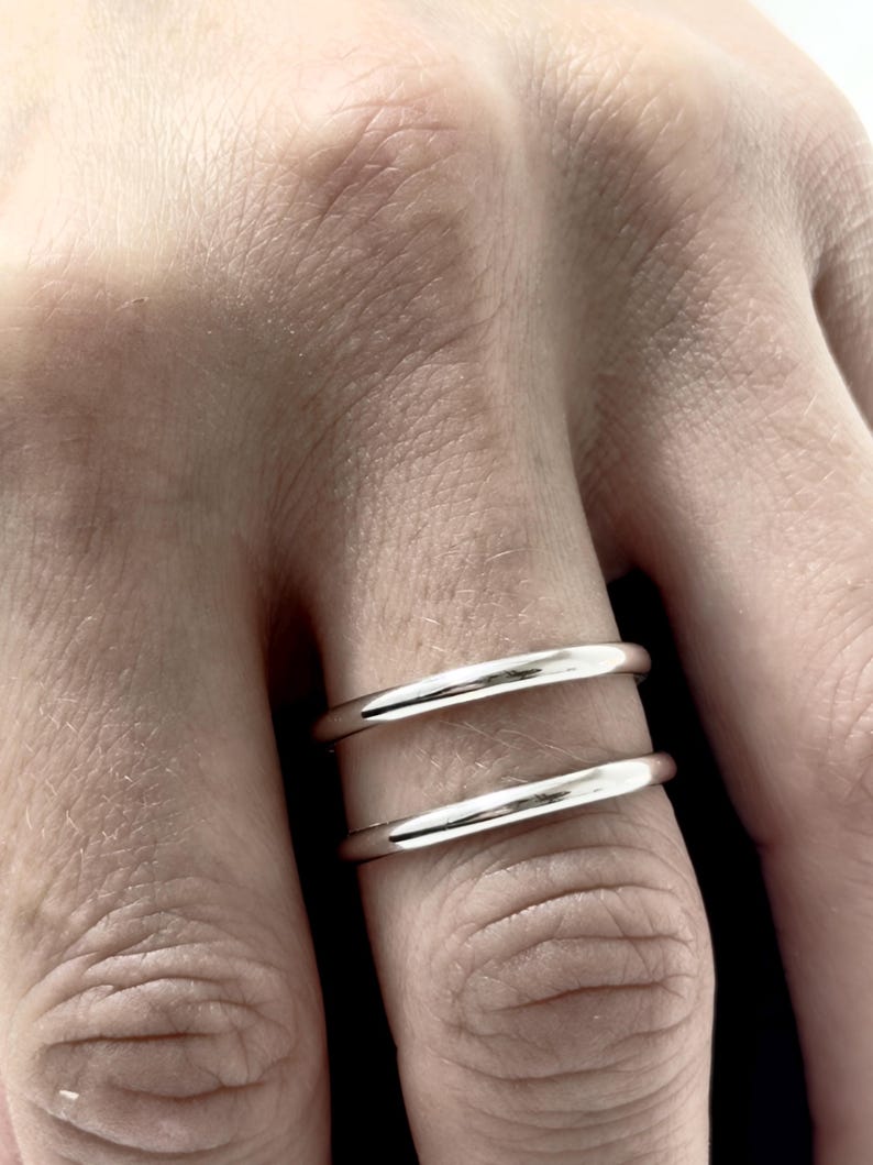 K&ouml;nnte beinhalten: Ein Set aus zwei glatten, polierten silberfarbenen Ringen, die an einem Finger getragen werden. Die Ringe haben ein schlichtes, abgerundetes Banddesign mit einer reflektierenden metallischen Oberfl&auml;che. Beide B&auml;nder sind in Form und Dicke identisch.