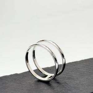 K&ouml;nnte beinhalten: Ein polierter Silberring mit doppeltem Banddesign. Die zwei d&uuml;nnen, glatten, kreisf&ouml;rmigen B&auml;nder sind an der Basis miteinander verbunden und bilden eine offene, parallele Struktur mit einer reflektierenden Metalloberfl&auml;che.