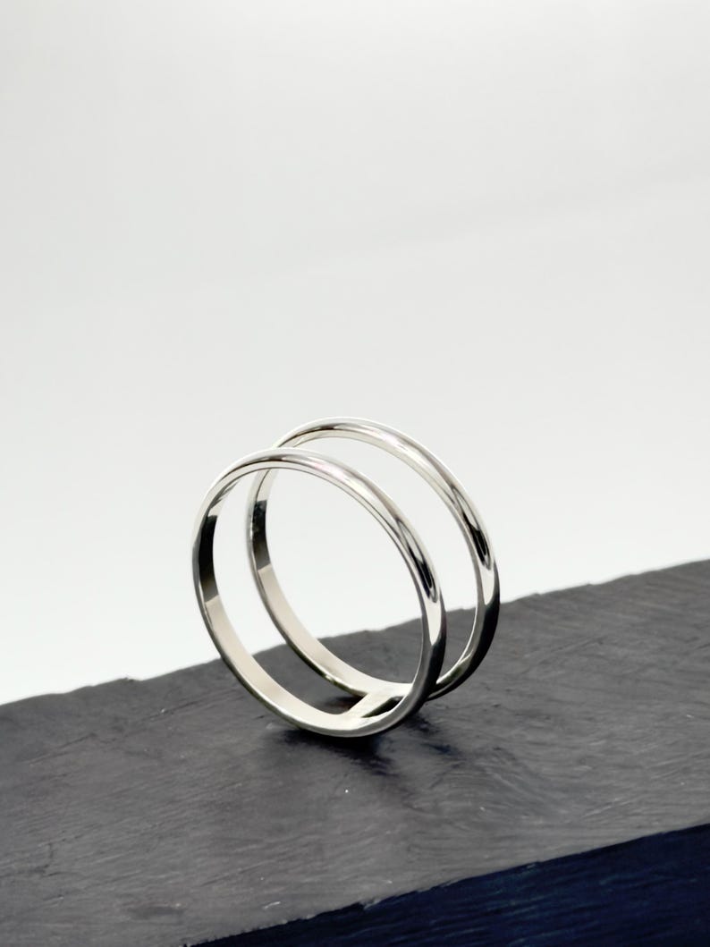 K&ouml;nnte beinhalten: Ein polierter Silberring mit doppeltem Banddesign. Die zwei d&uuml;nnen, glatten, kreisf&ouml;rmigen B&auml;nder sind an der Basis miteinander verbunden und bilden eine offene, parallele Struktur mit einer reflektierenden Metalloberfl&auml;che.