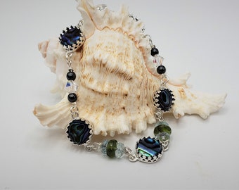 May include: A silver chain bracelet with abalone shell and black beads. The bracelet is displayed on a seashell.