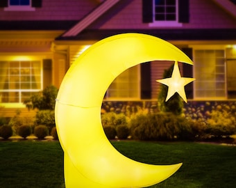 May include: An illuminated inflatable crescent moon and star decoration. The yellow moon arches over a star, both lit with a soft glow. The decoration is set on a green lawn in front of a house, creating a festive outdoor display.