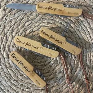 Op de afbeelding: Vier houten zakmessen met een leren band. Elk mes heeft de tekst "Bonne fête papa" en een hartje gegraveerd op het handvat. De messen zijn gepersonaliseerd met de namen "Elio & Robin", "Milo", "Elio" en "Eliana".