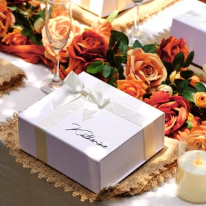 Könnte beinhalten: Weiße Geschenkboxen mit elfenbeinfarbenen Bändern und den Namen "Katherine" und "Olivia" in Schreibschrift werden auf einem Tisch präsentiert. Der Tisch ist mit orangefarbenen und roten Rosen, Sektgläsern und einer Kerze dekoriert.