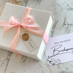 May include: A white gift box with a pink ribbon and a tag that says "Christina". The box is sitting on a marble surface next to a white card with the text "Will you be my Bridesmaid" and a floral design.