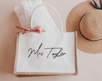 May include: A white tote bag with the text "Mrs Taylor" in black script, a pink bow, and a white towel. A straw sun hat with a black ribbon is also in the image. The bag has a natural-colored border.