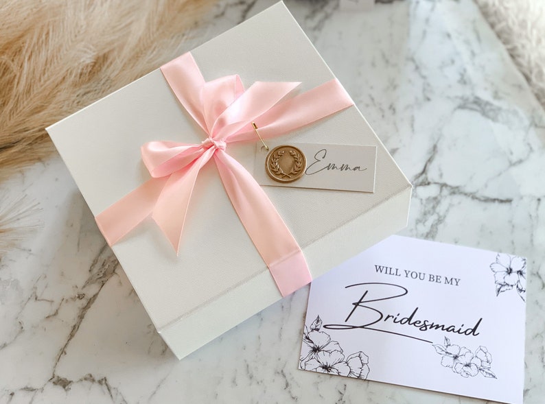 May include: A white gift box with a pink ribbon and a gold wax seal. The box has a tag that says "Emma". A card with the text "Will you be my Bridesmaid" is next to the box.