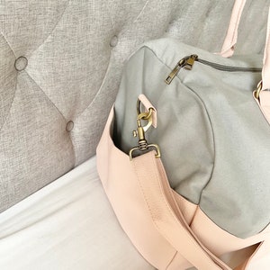May include: A grey and pink duffel bag with a shoulder strap and a zip closure. The bag has a light pink faux leather base and accents, with gold-coloured hardware. The bag is on a white surface.
