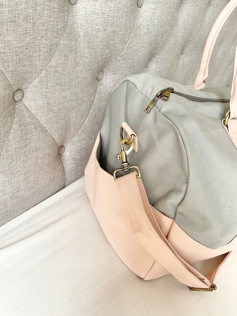 May include: A grey and pink duffel bag with a shoulder strap and a zip closure. The bag has a light pink faux leather base and accents, with gold-coloured hardware. The bag is on a white surface.