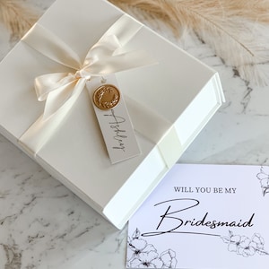 May include: White gift box with a white satin ribbon and a gold wax seal. The box has a tag that says "Ashley" on it. A white card with black text says "Will you be my Bridesmaid?" with floral designs.