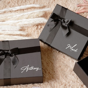 May include: Two dark gray gift boxes with black satin ribbons and bows. One box has the name "Anthony" in white script, and the other has "Mark". The boxes are on a beige rug.