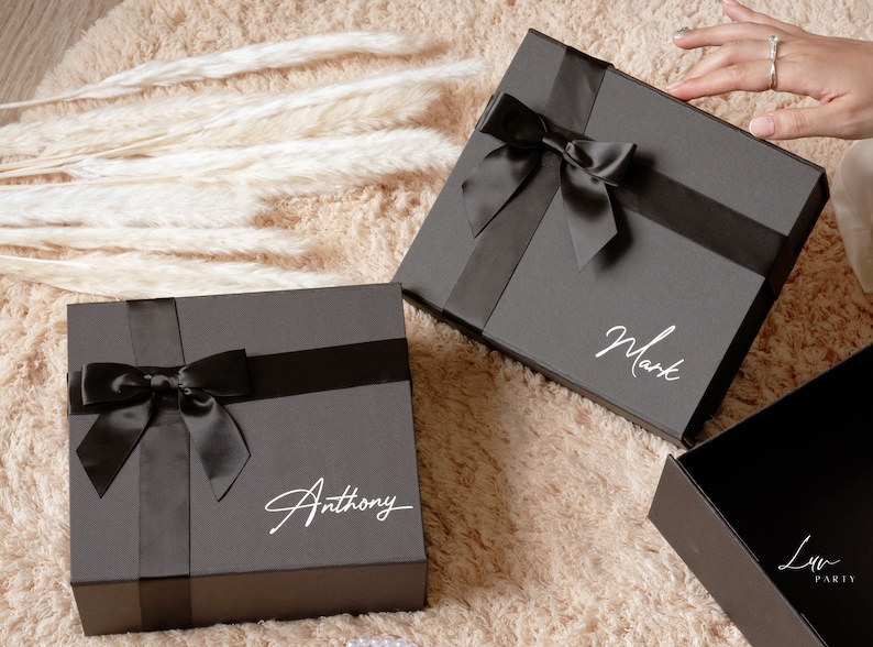 May include: Two dark gray gift boxes with black satin ribbons and bows. One box has the name "Anthony" in white script, and the other has "Mark". The boxes are on a beige rug.