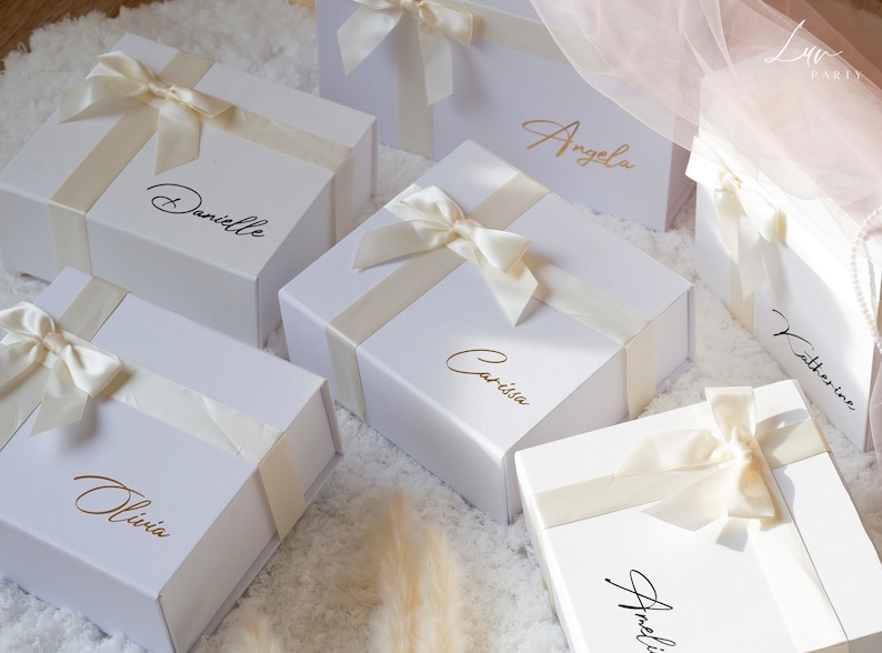May include: White gift boxes with ivory satin ribbons and bows. Each box is personalized with a name in either gold or black script. The boxes are arranged on a white, textured surface, suggesting a celebratory theme.