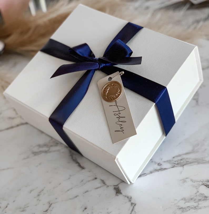 May include: A white gift box with a navy blue ribbon and bow. A small white tag with the name "Ashley" is attached to the ribbon. The tag has a gold wax seal with a wreath design.