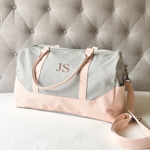 May include: A grey and pink duffel bag, featuring the initials "JS". The bag has a light grey canvas body with pink faux leather accents on the base and handles. It includes a detachable shoulder strap.