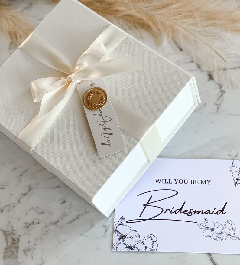 May include: White gift box with a white satin ribbon and a gold wax seal. The box has a tag that says "Ashley" on it. A white card with black text says "Will you be my Bridesmaid?" with floral designs.