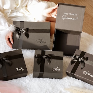May include: A collection of dark gray gift boxes with black ribbon and bows. Each box has a name in a cursive font. One box is open, revealing the text "WILL YOU BE MY Groomsman?"