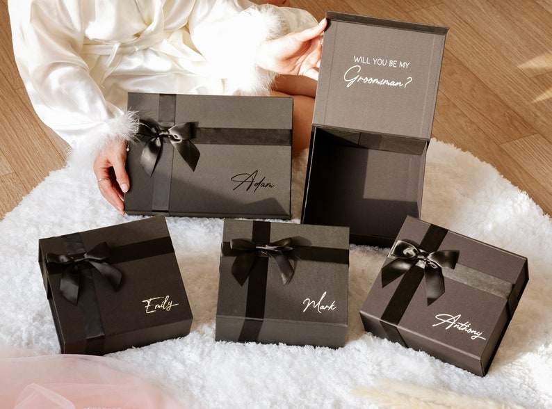 May include: A collection of dark gray gift boxes with black ribbon and bows. Each box has a name in a cursive font. One box is open, revealing the text "WILL YOU BE MY Groomsman?"