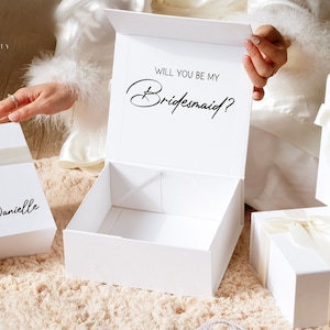 May include: White gift boxes with ivory ribbon and the text "Will you be my Bridesmaid?" in black script. One box is open, revealing an empty interior. Other boxes are closed, with names written on them.