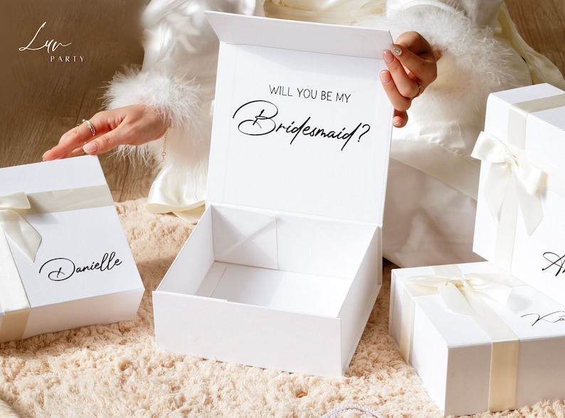 May include: White gift boxes with ivory ribbon and the text "Will you be my Bridesmaid?" in black script. One box is open, revealing an empty interior. Other boxes are closed, with names written on them.