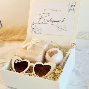 May include: A white gift box with a gold lid, filled with a white bath bomb, a small white box with the name "Ashley" on it, a pair of white heart-shaped sunglasses with the word "Babe" on them, and a white pen with a gold top. The lid of the box has a card that says "Will you be my Bridesmaid" with a floral design.