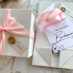May include: A white gift box with a pink ribbon and a tag that says "Emma". The box is next to a white box with a pink ribbon and a card that says "Will you be my Bridesmaid?" with a floral design.